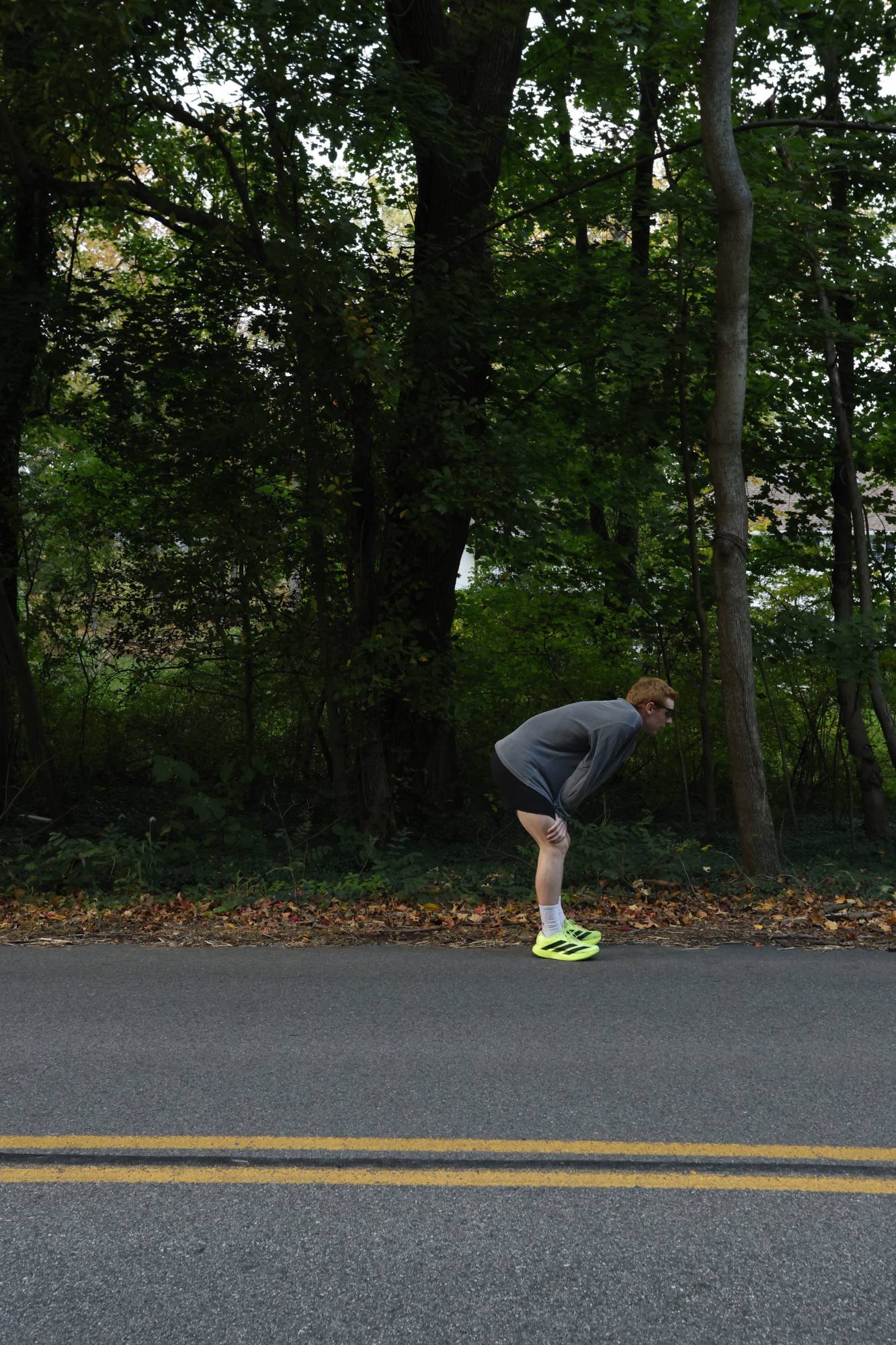 A runner in a grey long-sleeve and neon shoes, hands on knees at the edge of a tree-lined road